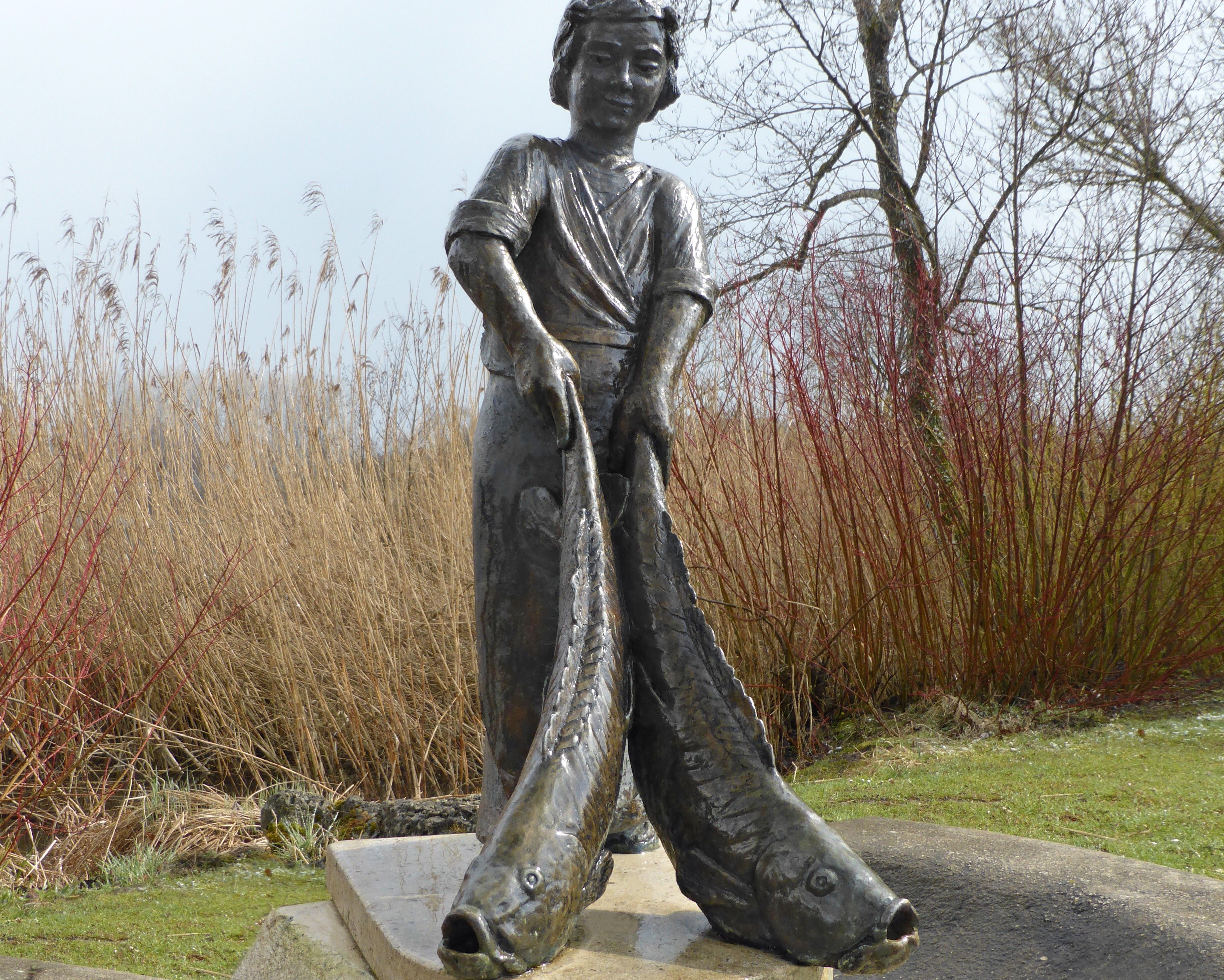 Eine Bronzestatue auf einem Sockel zeigt einen Fischerbub, der in beiden Händen einen Fisch hält. 