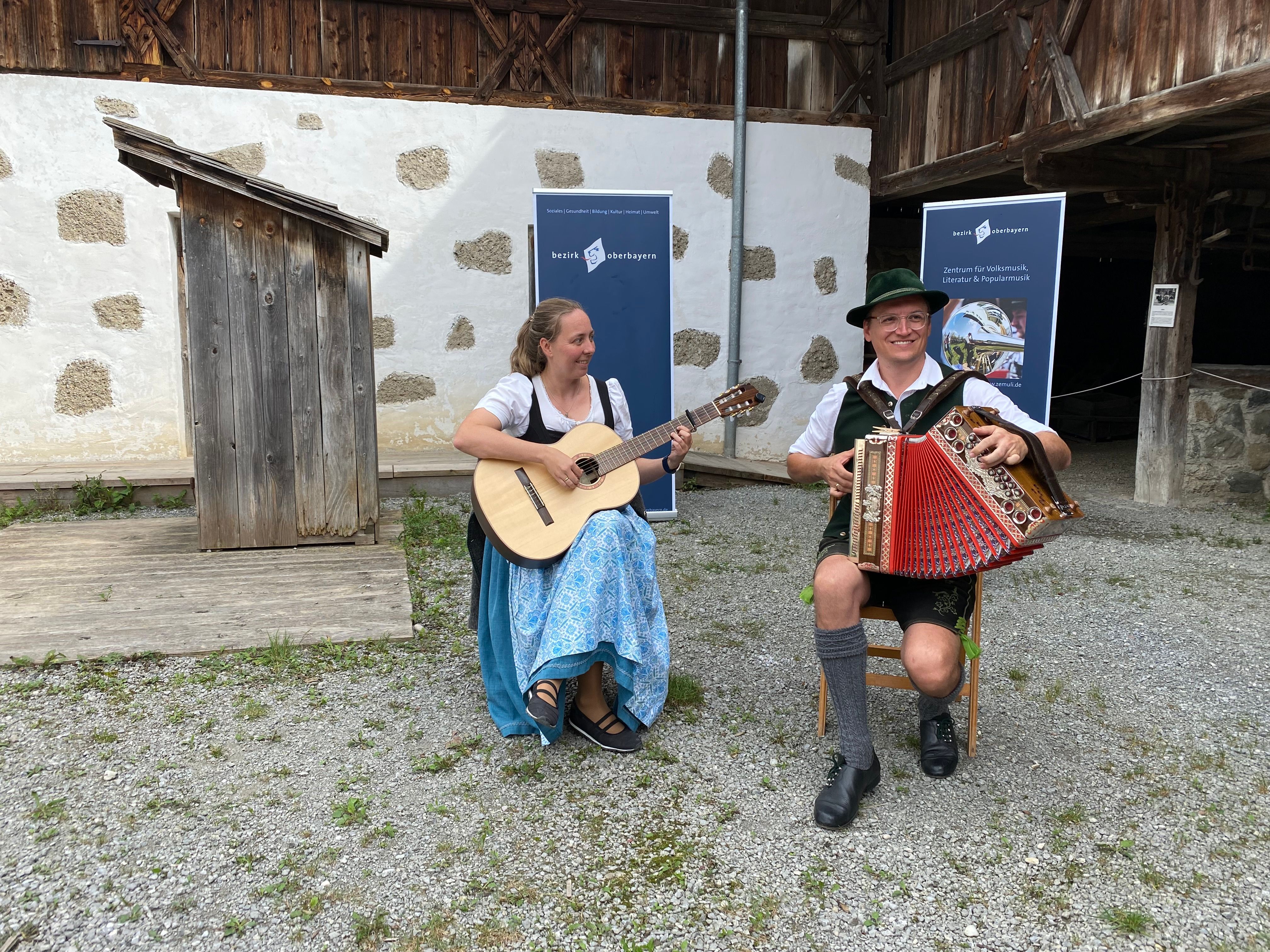 Ein Mann in Tracht spielt eine steirische Harmonika, eine Frau im Dirndlkleid spielt Gitarre und sitzen beide auf einem Stuhl vor einem alten Gebäude.