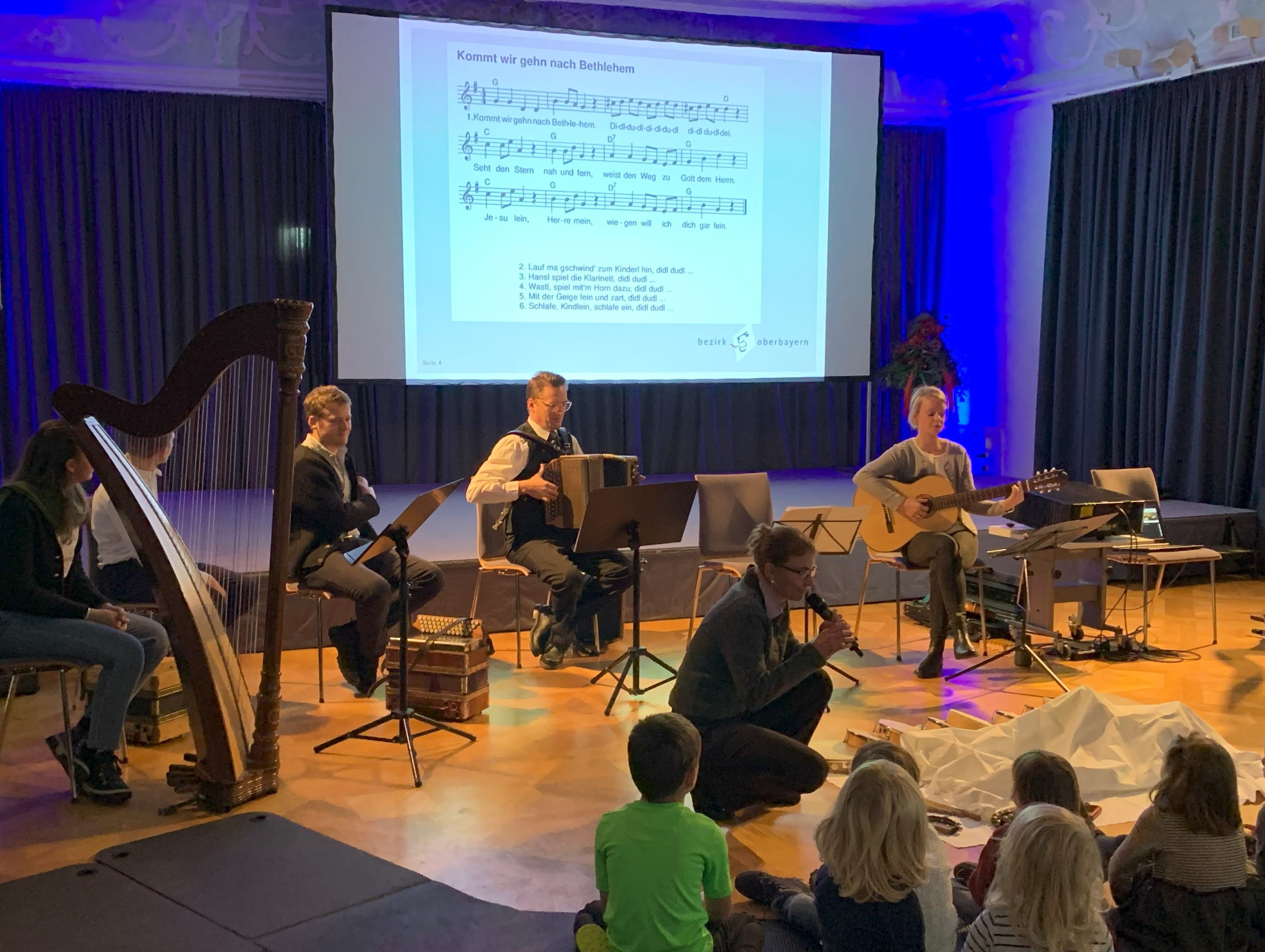 Mitarbeiterinnen und Mitarbeiter des ZeMuLi singen und musizieren mit Kindern, die am Boden auf Weichbodenmatte sitzen.